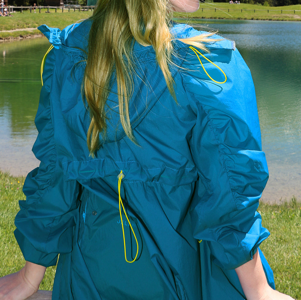 Mont Blanc ultralight long windproof jacket in bright blue with adjustable waist and yellow drawstrings worn outdoors near a lake