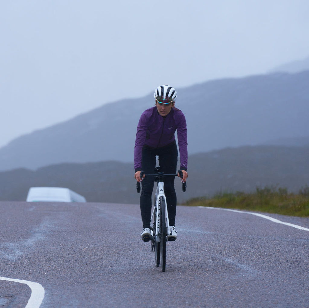 Cyclist wearing purple 3L Core Rain Jacket riding on wet road in cloudy weather